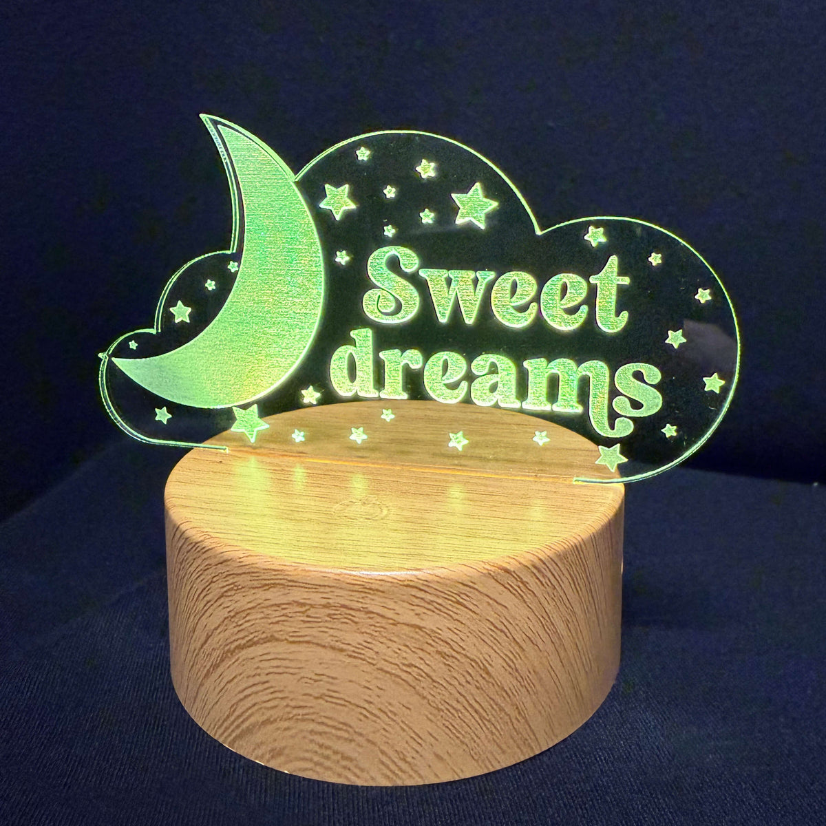 A wooden night light with a clear acrylic sign that reads 'Sweet dreams' in green, decorated with a crescent moon and stars, displayed on a round wooden base against a dark blue background.