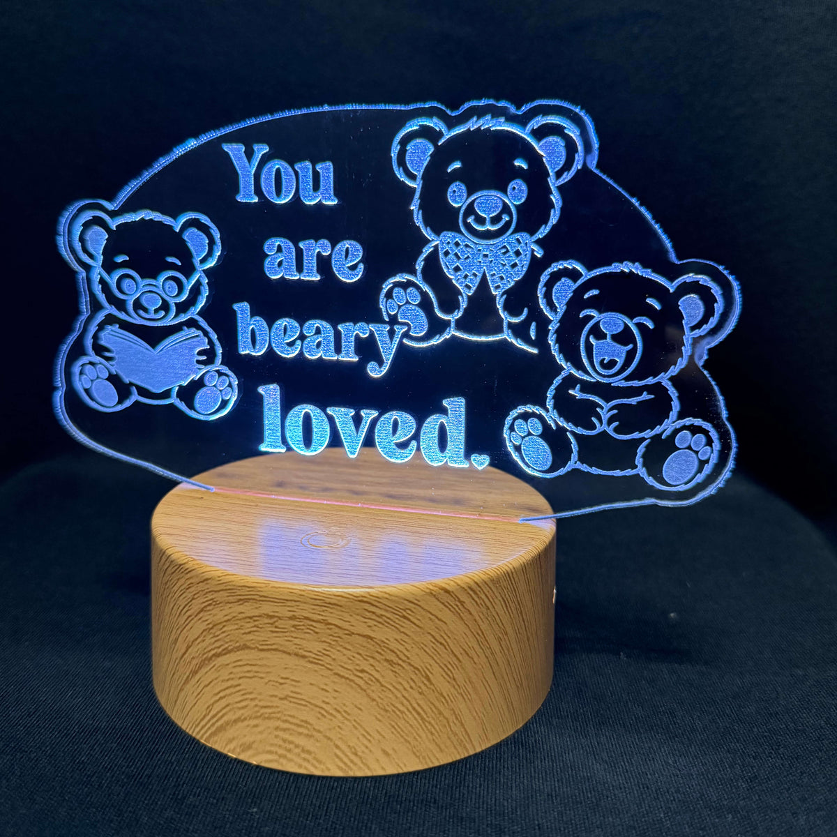 A wooden cylindrical night light base with a glowing blue acrylic plaque above it, featuring teddy bear illustrations and the text 'You are beary loved.' on a dark background.